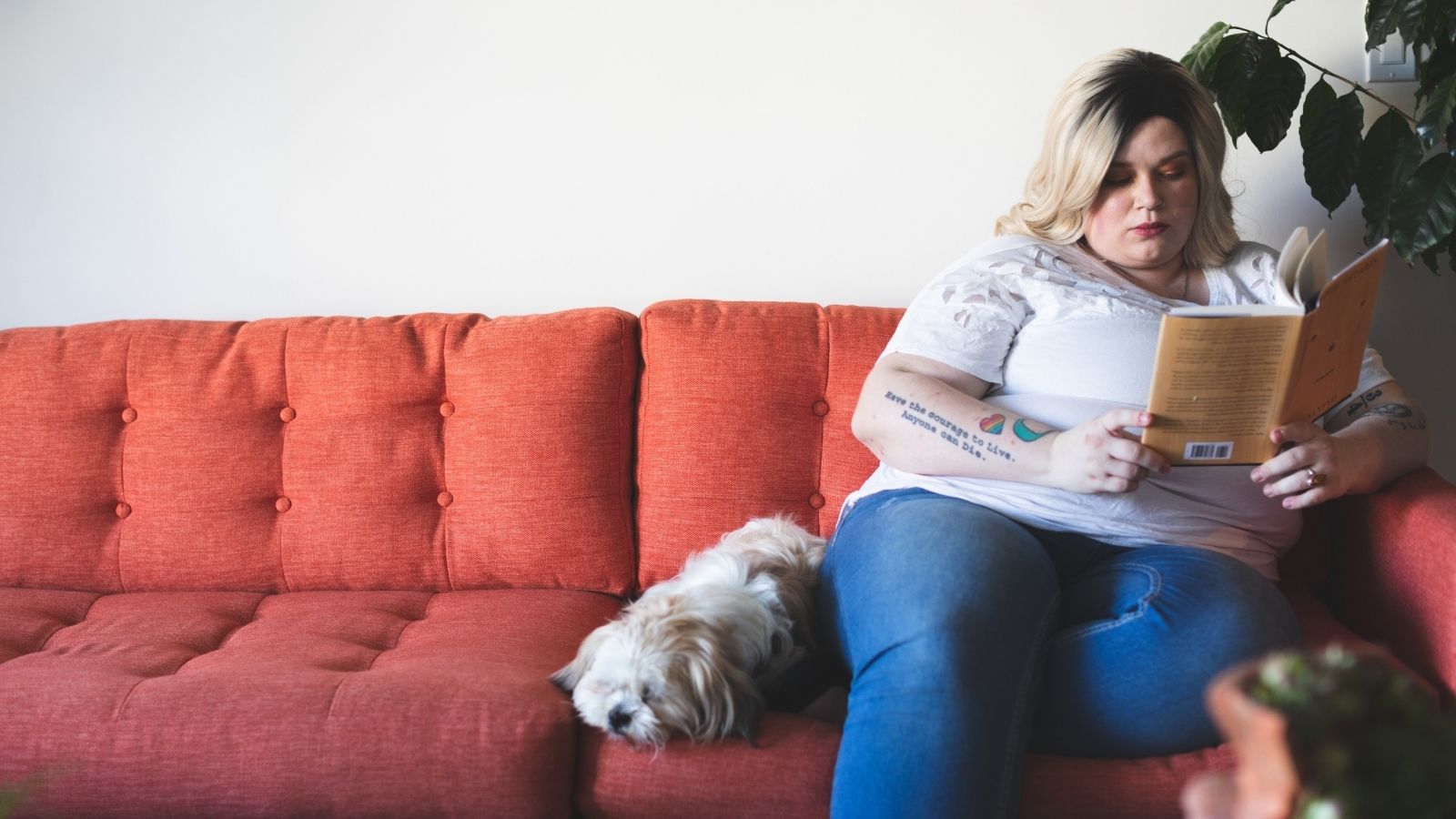 An obese woman sits on the couch with her dog reading a book