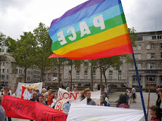 Kundgebungsteilnehmerinnen mit Transparenten, »Atombomben raus!«, und Regenbogen-Friedensfahne »PACE«.
