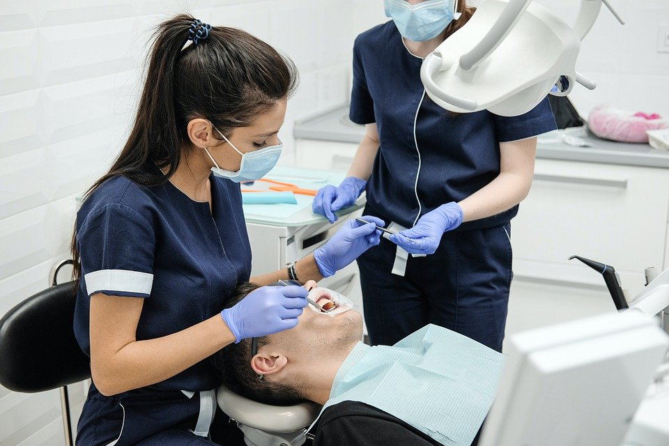 A female dentist wearing a medical mask performs a dental procedure on a man reclined in the dental chair