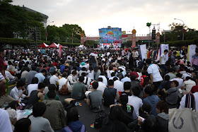 large crowd sitting on the ground with a colorful stage in the distance