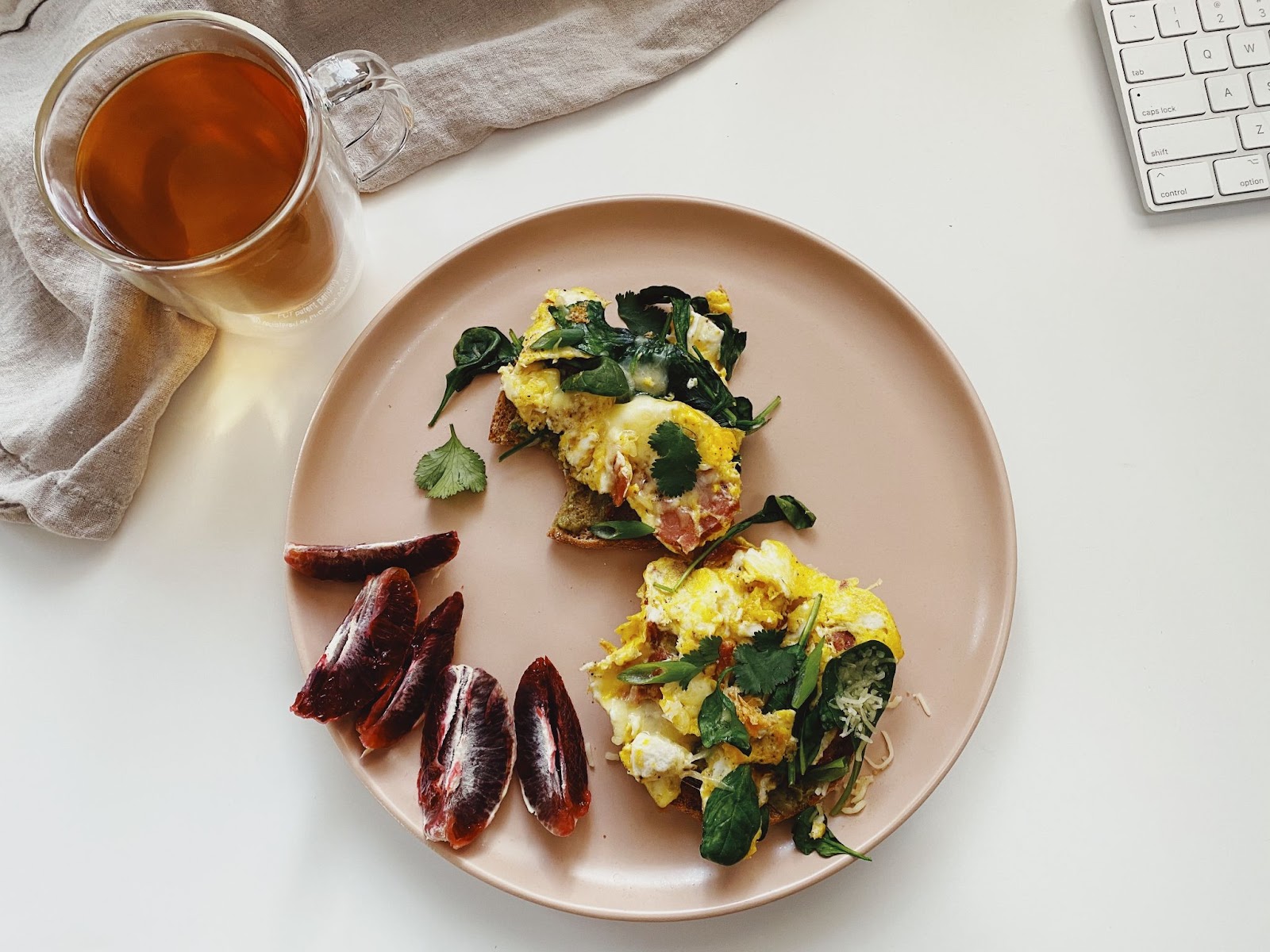 Pink plate with avocado toast on English muffin, topped with scrambled eggs, spinach, and turkey bacon. A cup of tea on the side.