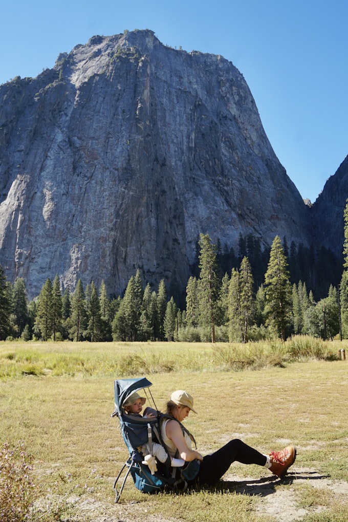 A person is sitting on the ground at the base of a tall rock mountain while wearing a kid carrier with a toddler in it.