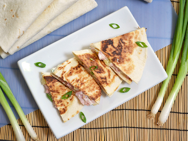 Top view of Hawaiian Ham Quesadillas sliced into four slices on plate, whole green onion on the side 