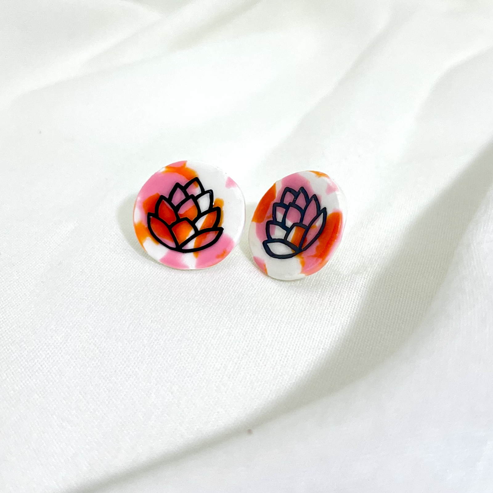 A pair of round earrings featuring a dark outline of a hop cone, on a marbled background of orange, pink and white. 