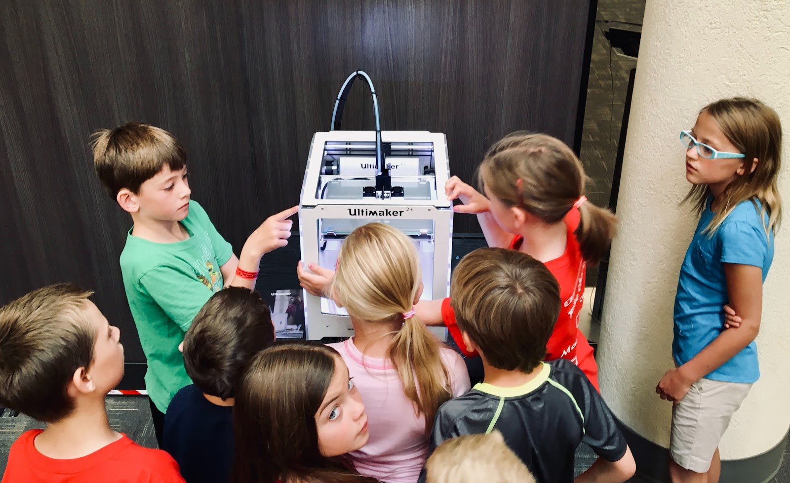 About ten elementary students are gathered around the 3-D printer in their classroom, observing it in action.