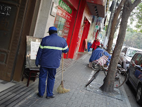street cleaner with a handmade broom reading a sign in Shanghai