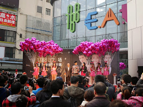 3 people and 6 young women wearing pink dresses and bunny ears while holding large bunches of heart-shaped balloons stand on a stage for an outdoor promotion