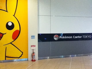 Photo of a wall. On the left is half of a smiling pikachu art from floor to ceiling. Next to the image of pikachu is a black bar that says "Pokemon Center Tokyo" with a pokeball icon