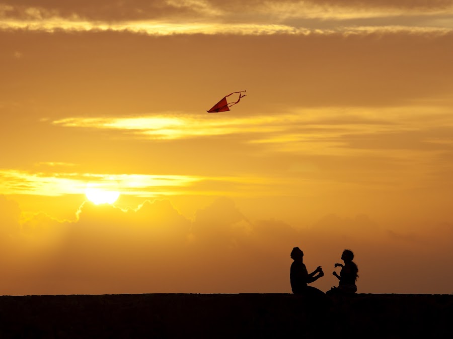 A couple sit together on a wall overlooking the Indian Ocean, flying a kite as the sun sets