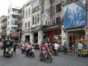 motorbikes, an auto-rickshaw, a food stall, pedestrians and an advertisement for PEAK NBA products with the words "I can play" on Xinhua Road in Haikou