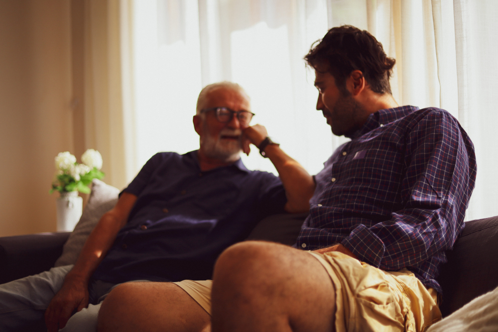 Boomer dad and GenX son sitting on couch at home and talking.