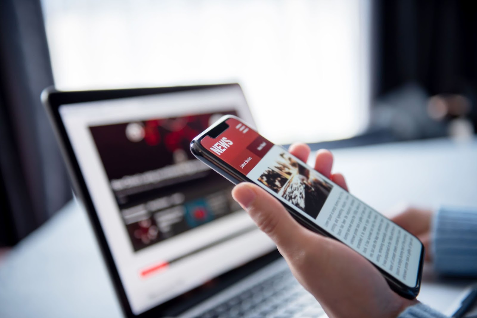 a hand holding a mobile phone with news article on screen and an opened laptop in the background