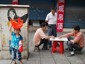 boy smiling and holding a small plastic stool over his head near men playing xiangqi (Chinese chess)