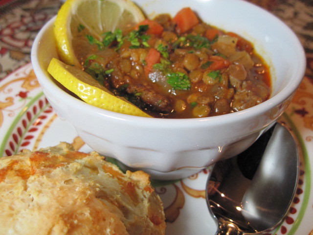 A bowl of Lentil Stew with lemon slices, a spoon, and a biscuit.