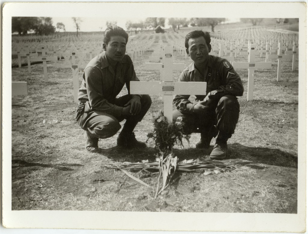 Two Japanese American soldiers crouch down next to a grave in a military cemetery. There are flowers on the ground next to the grave.