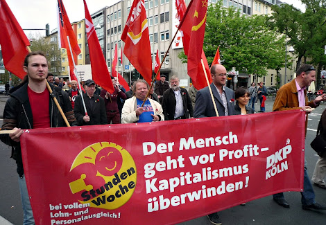 Demonstranten mit DKP-Fahnen und Transparent: »Der Mensch geht vor Profit – Kapitalismus überwinden! DKP Köln«.