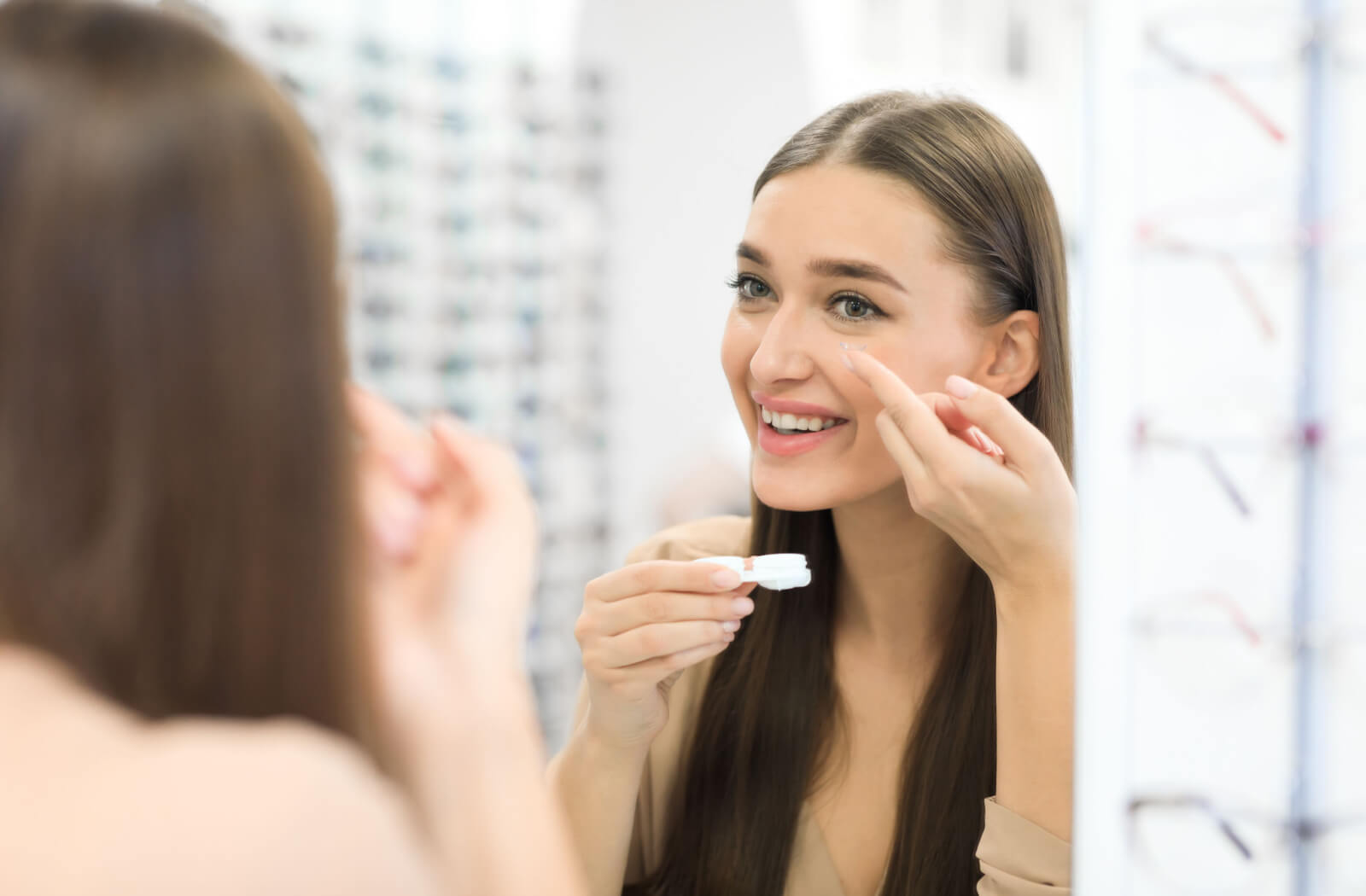 A woman with long hair looking in the mirror and holding a contact lens container in her right hand and inserting a contact lens with her left index finger.