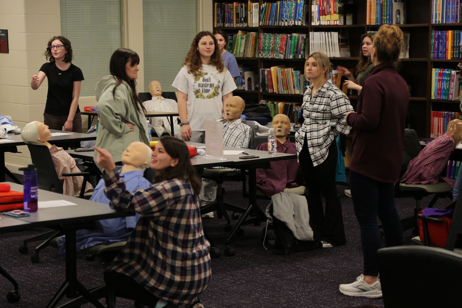 photo of instructor and 7 students at the tables in the ETMC each standing near a CPR dummy that is upright in a chair