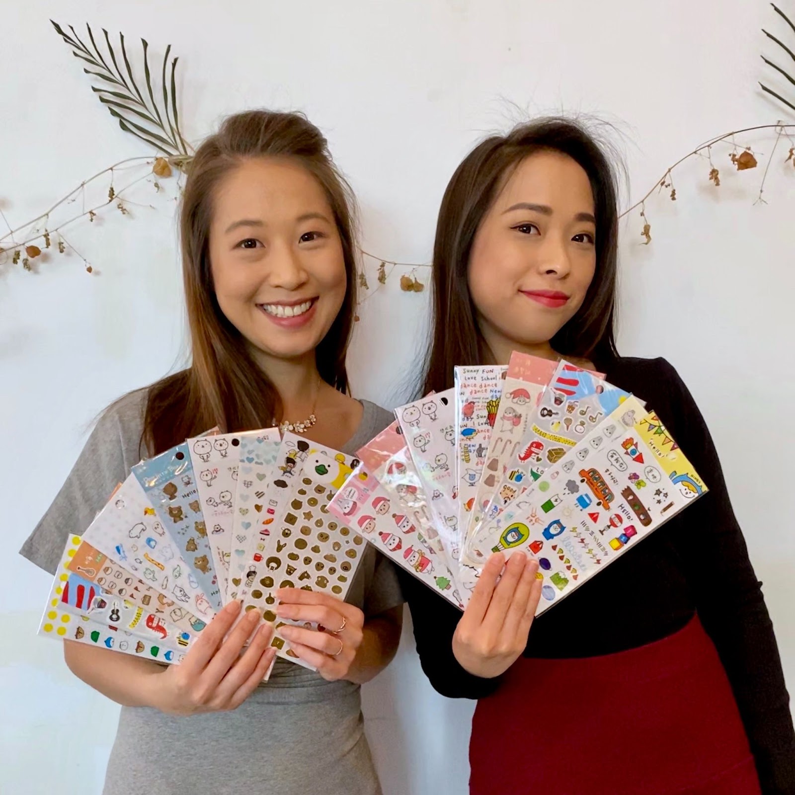 SmileABCs Sticky Rice Sisters–The image shows Koyun and Kochin smiling side by side, each displaying several packs of stickers.