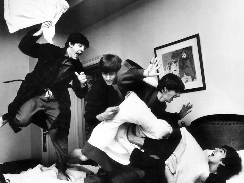  Temos os quatro integrantes da banda Beatles em cima da cama, em uma guerra de travesseiros. 