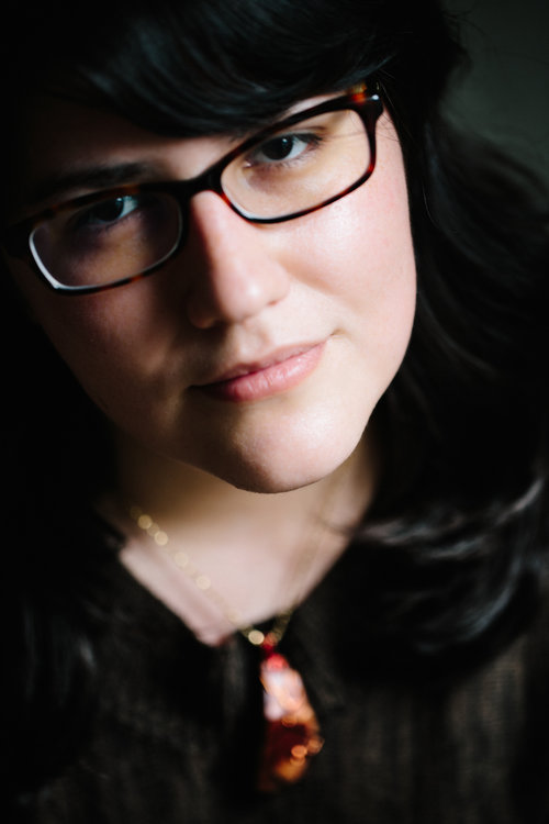 A bespectacled brunette wearing a large stone necklace tilts her head at the camera
