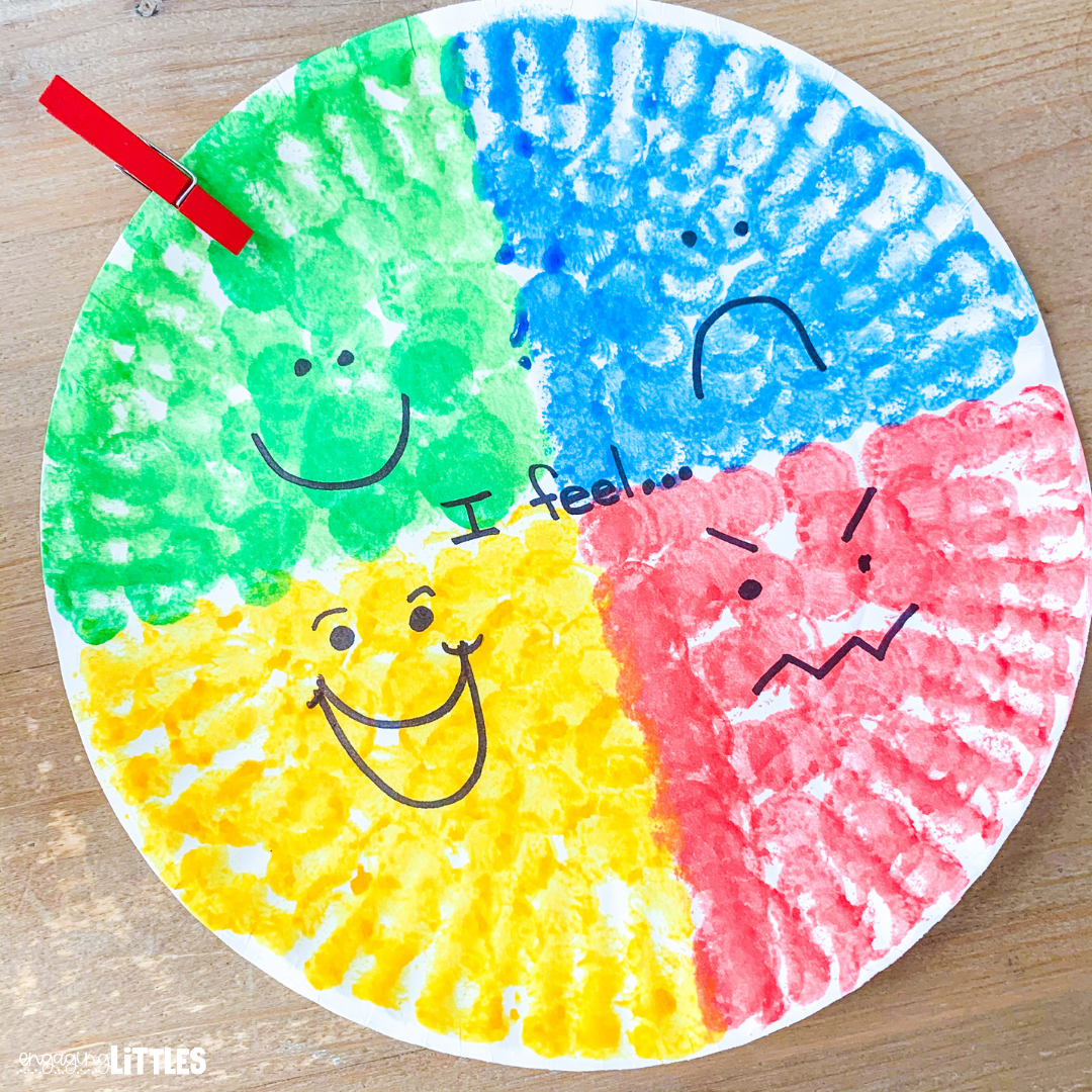 A paper plate divided into four sections - top left quadrant is green with a happy face drawn on it; top right quadrant is blue with a sad face; bottom right is red with an angry face; bottom left is yellow with an excited face. In the center of the plate are the words: "I feel...". There is a red clothespin attached to the top left green quadrant.