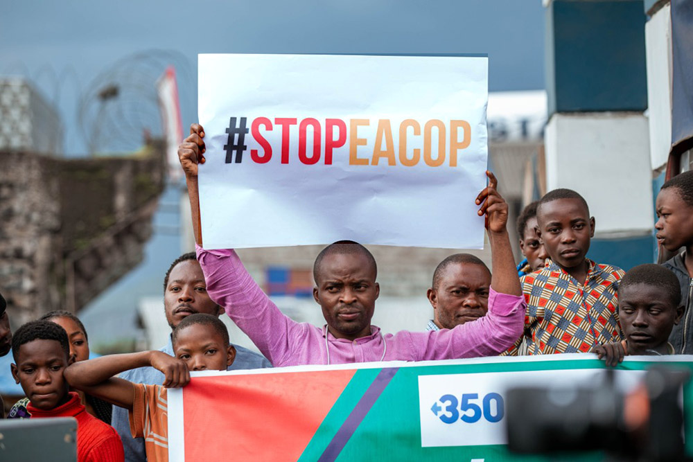 A man holds up a large piece of card withe message '#STOP EACOP', with young boys to each side of him