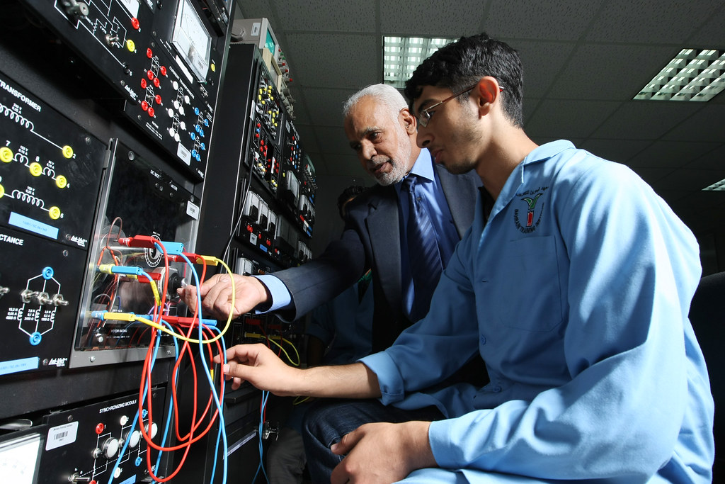 An older gentleman in a suit mentors a young student intern in a lab coat as they explore wiring and connections on a large circuit board.