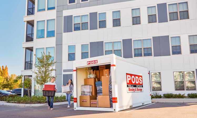 A couple is carrying a moving box, a cooler, and a plastic bin to their PODS portable moving and storage container in the parking lot of their apartment complex. The container is loaded with many moving boxes, some furniture, and a mattress.