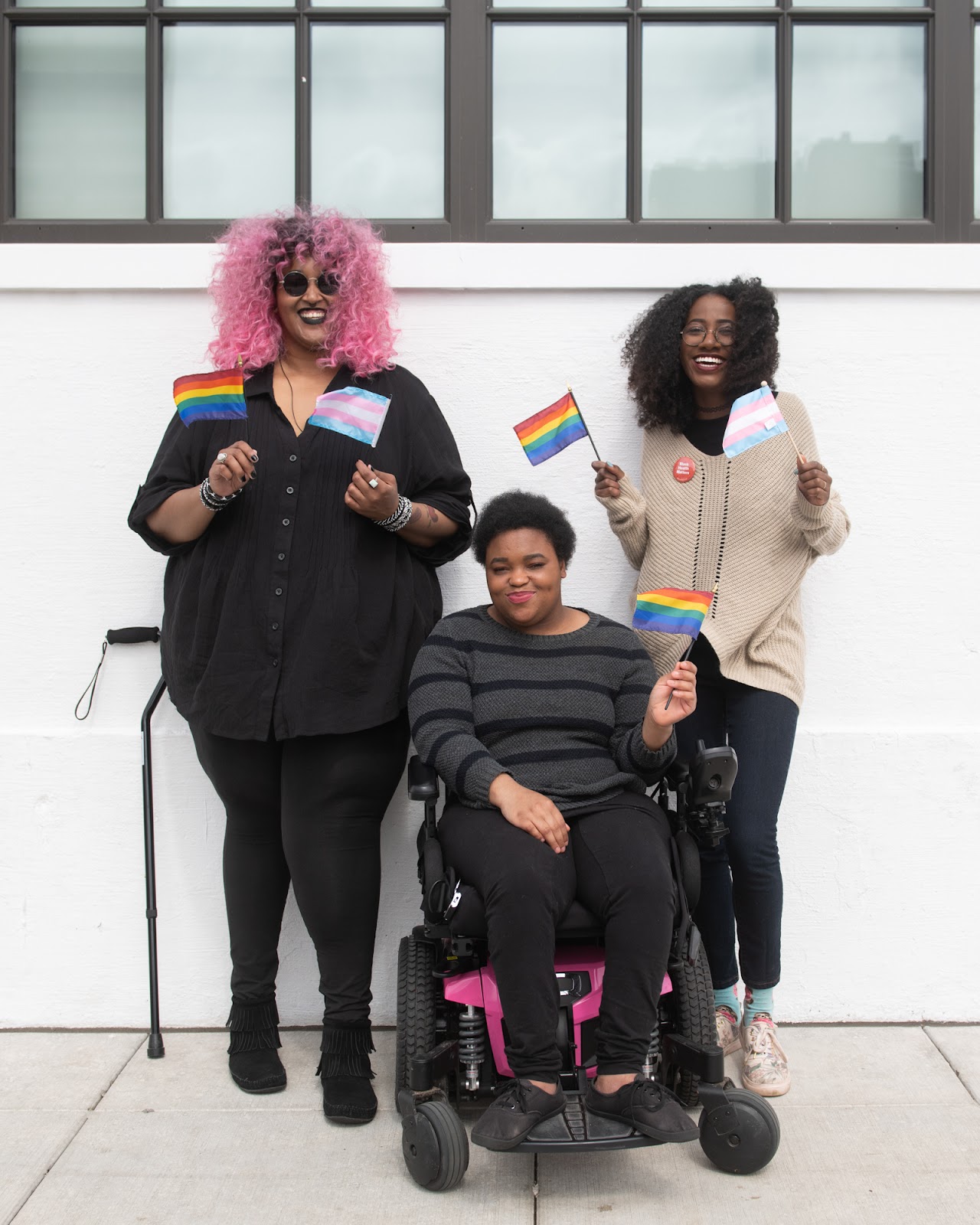 Three Black and disabled folx smile and hold mini flags. On the left, a non-binary person holds both a rainbow pride flag and a transgender pride flag, while a cane rests behind her. In the middle, a non-binary person waves the rainbow flag while in their power wheelchair. On the right, a femme waves both a rainbow and transgender pride flag. (via Disabled and Here image collection)