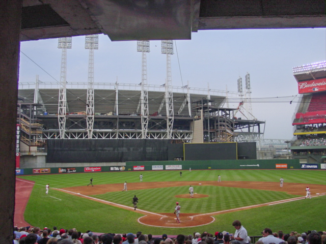 The outfield of a ballpark is shadowed by a large unfinished stadium in the outfield. 