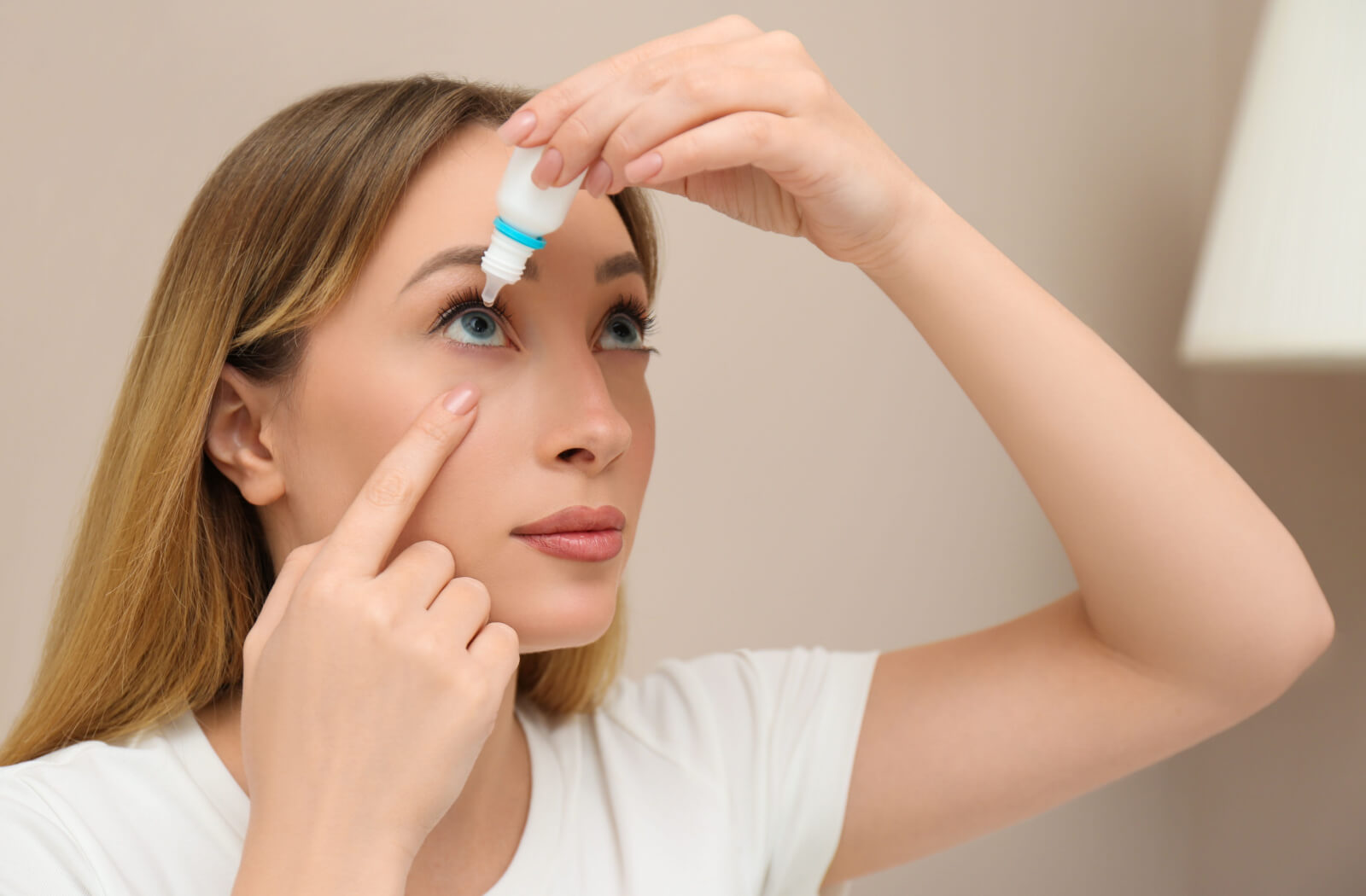 A young woman pulling her right lower eyelid down to apply eye drops while standing against a beige background.