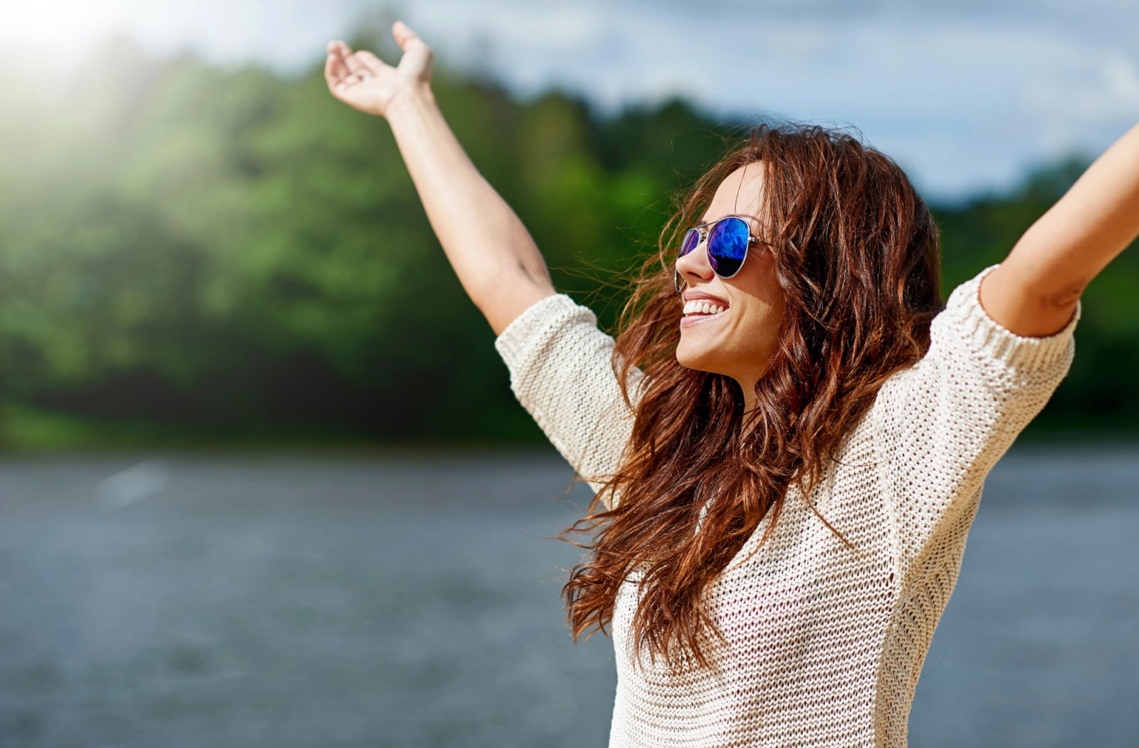 A woman wearing polarized sunglasses, smiling and holding her arms up in the air while she is outside.