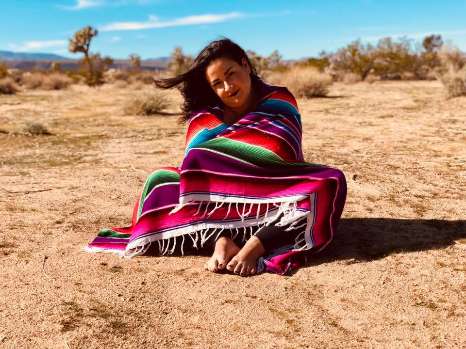 Melissa sits on the ground of the desert wrapped in a sarape blanket and smiles in the middle of at the camera.