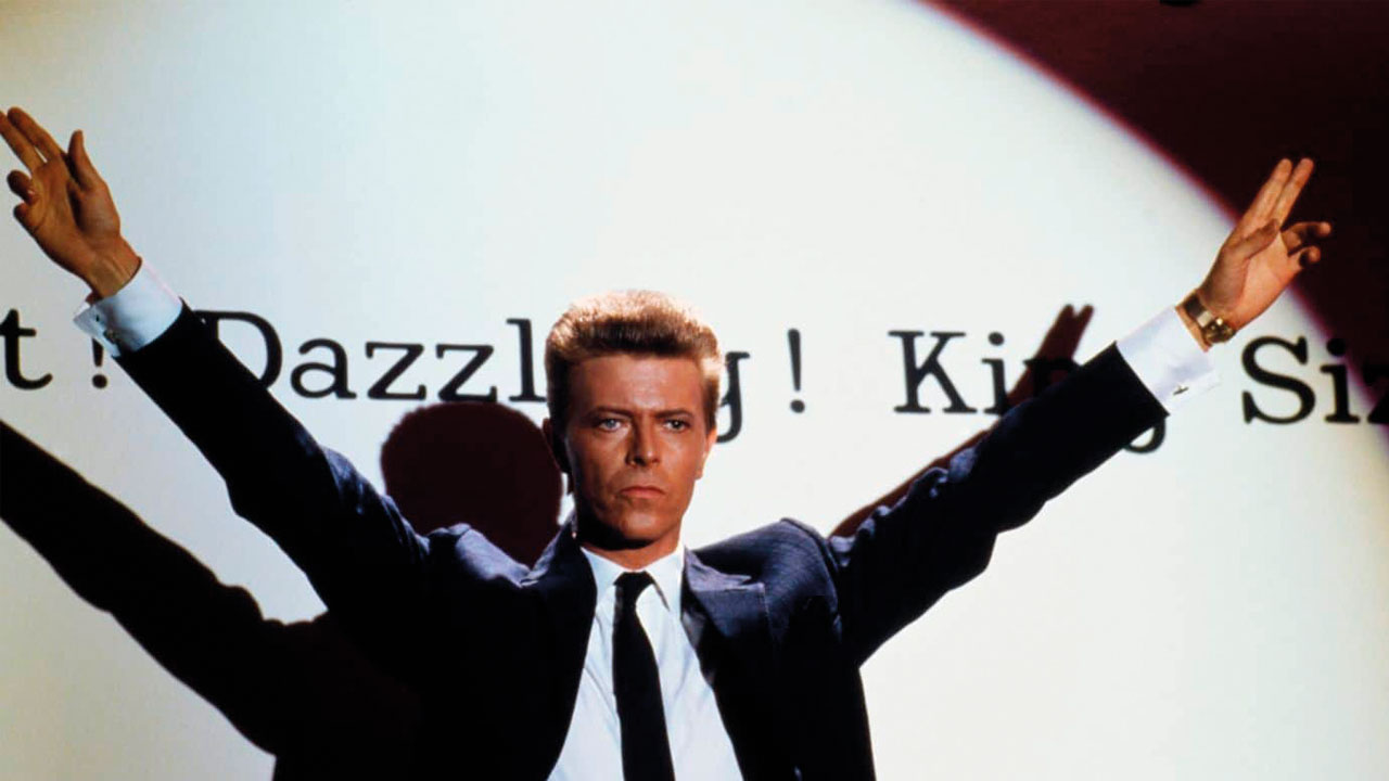 Bowie stands in the centre of the image, words like 'dazzling' partially obscured behind his head on the white wall he is in front of. He stands looking out, arms raised in a V shape above his head. His hair is short, and he is wearing a suit.