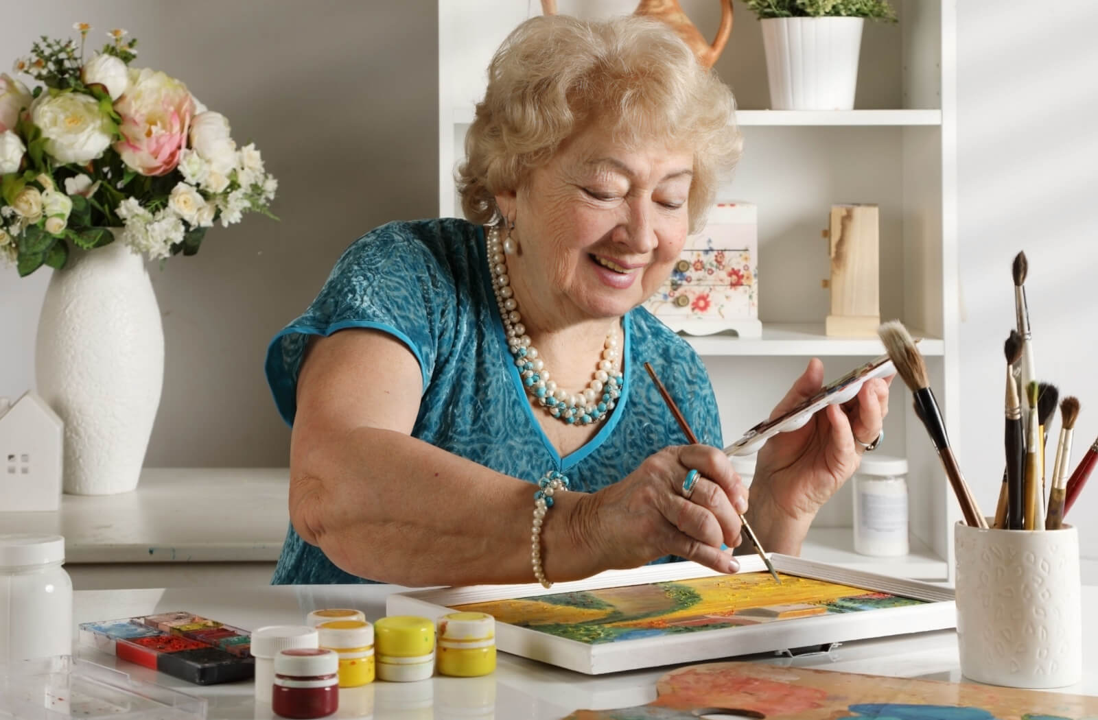 A senior woman is sitting at the table coloring a painting on a canvas at home with a small bottle of paint colors,and  paint brushes