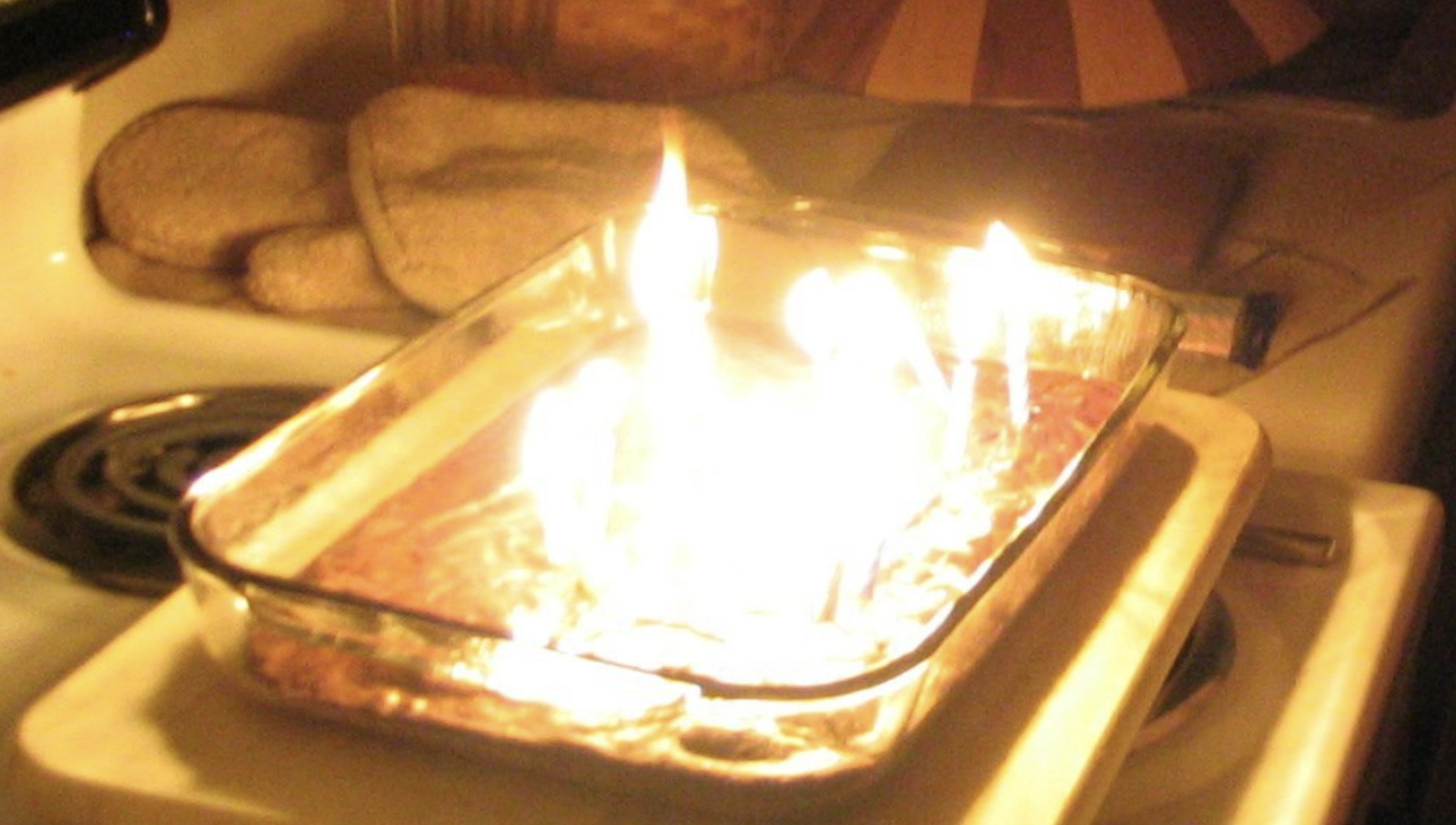 The photo is of a rectangular glass cake pan with contents that appear to be on fire with the fire being the only source of light illuminating the cake pan in yellow light, sitting on an old coil burner electric stove top next to a counter with oven mitts.