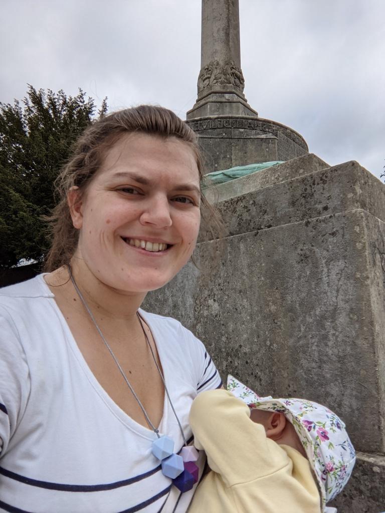 Fiona, the author, sitting on stone steps by a war memorial in a white, navy-striped t-shirt, breastfeeding her daughter who is wearing a yellow cardigan and floppy, floral sun hat.