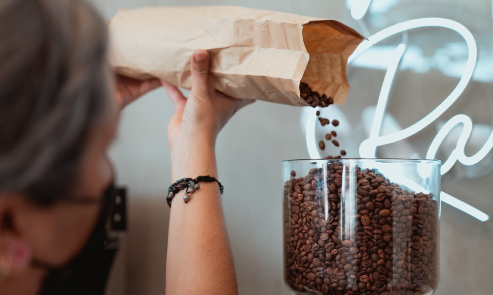 Caucasian female pouring roasted coffee beans from brown kraft paper bag into coffee grinder hopper with neon sign in background. 