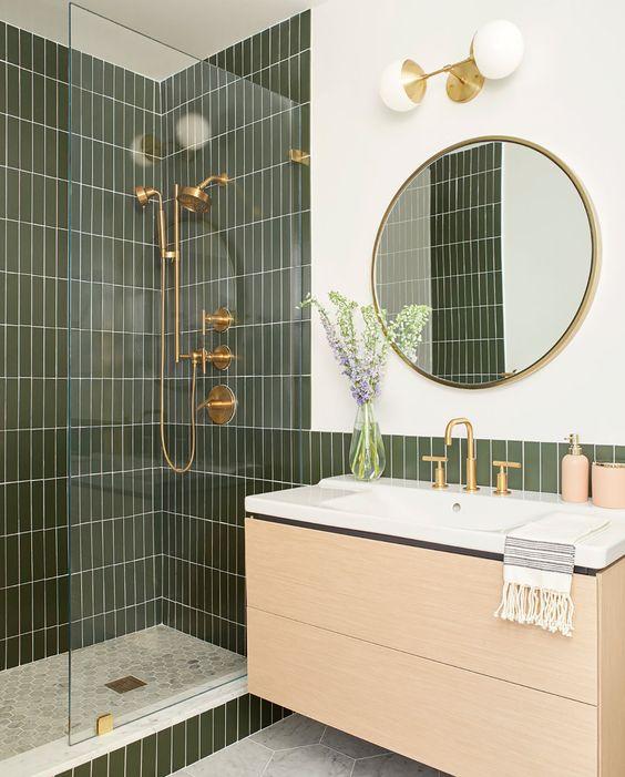 Banheiro com revestimento subway tiles verde musgo na área do box e metade da parede da pia, piso hexagonal em formato pequeno em cor neutra no box e hexagonal em formato grande em tom cinza fora do box, armário amadeirado claro e pia branca com louças, chuveiros e moldura do espelho dourado.