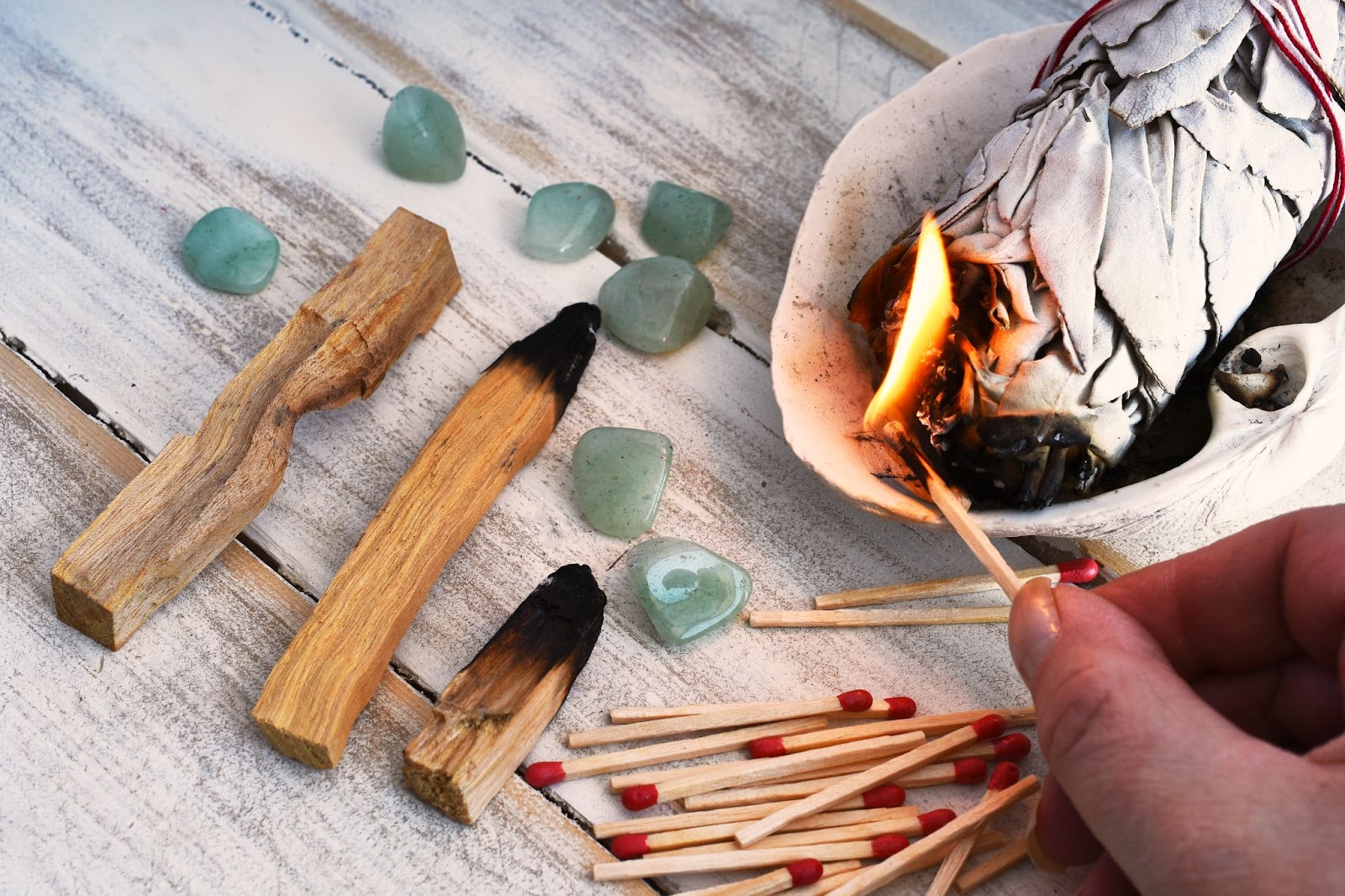 Green crystals next to a stick of sage and sticks of palo santo with a hand holding a lit flame.