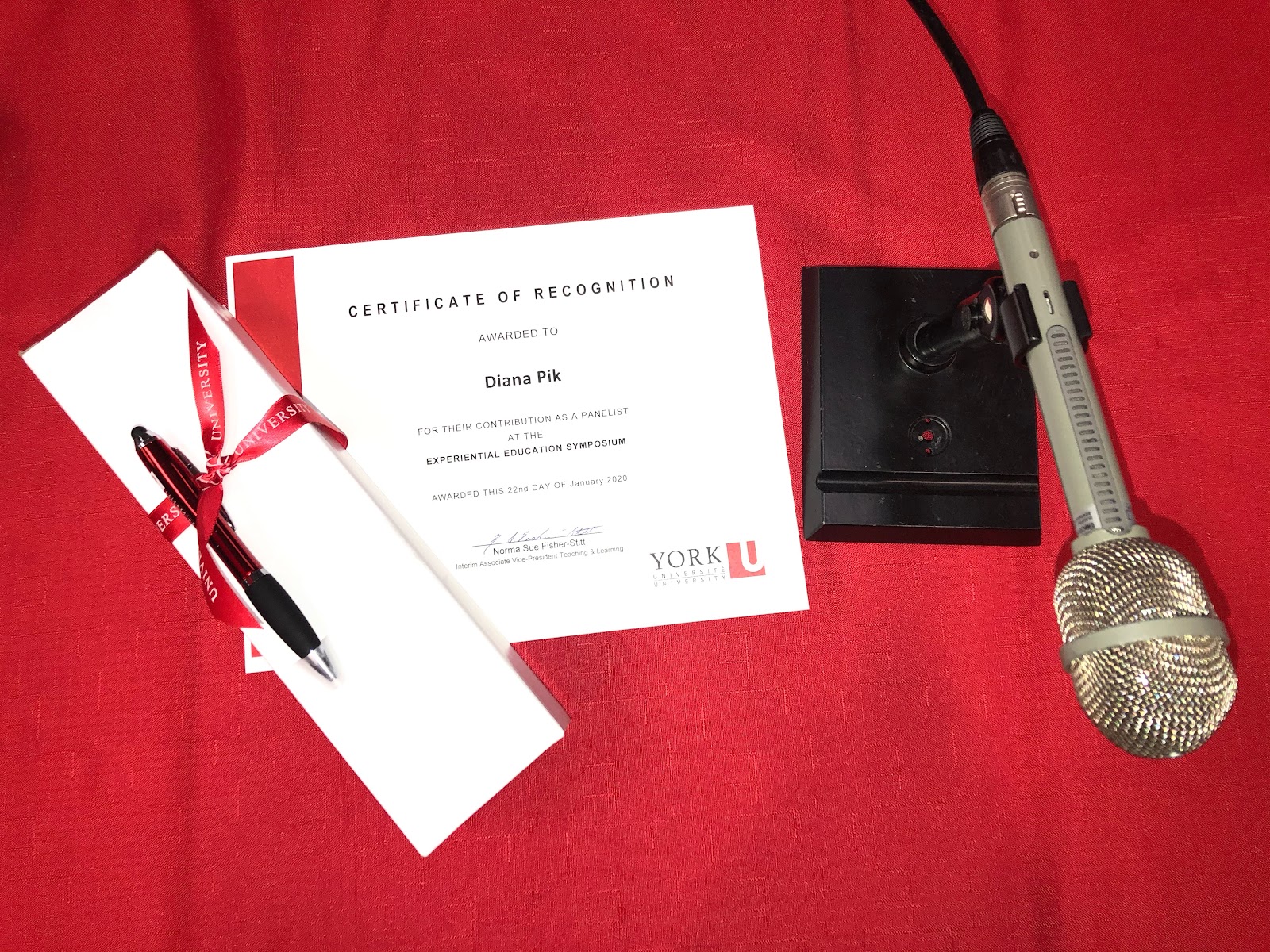 Four objects on a table. left to right the objects are a box with a pen attached to it, a certificate of recognition, and a microphone. 
