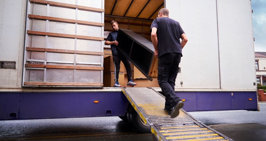 Two men are unloading a tall bookshelf from a moving truck. One is walking down the built-in ramp, and the other is about to step on the ramp.