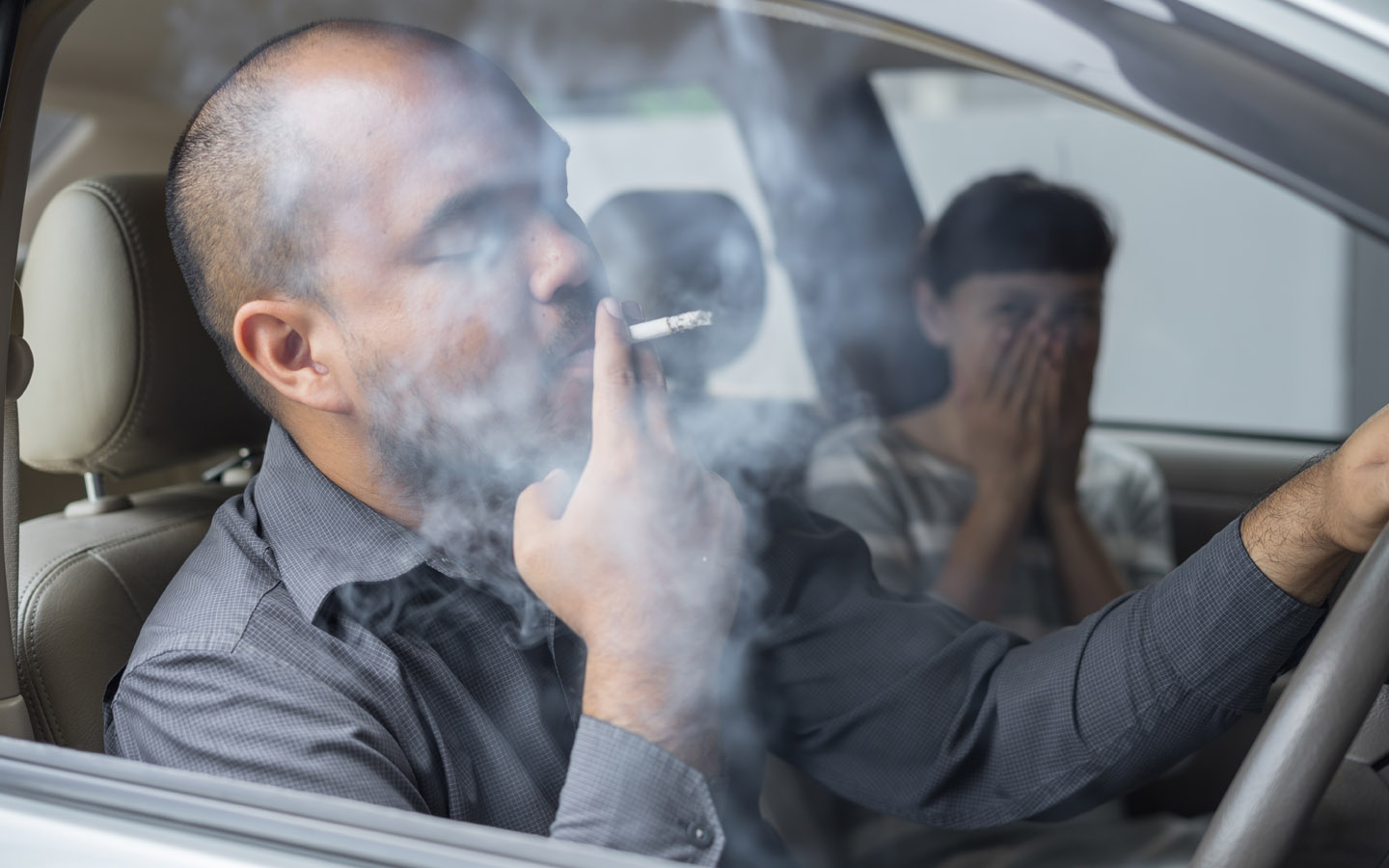 Мужчина курит сигарету в машине, в то время как пассажирка испытывает отвращение к дыму