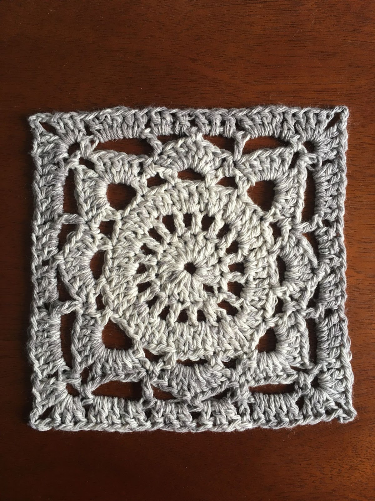A grey crocheted square, designed in an intricate, snowflake-like pattern.
