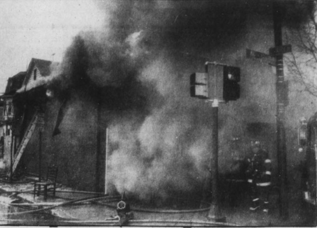 Black and white image of a one story brick building with lots of smoke,, a chair and hoses on sidewalk.
