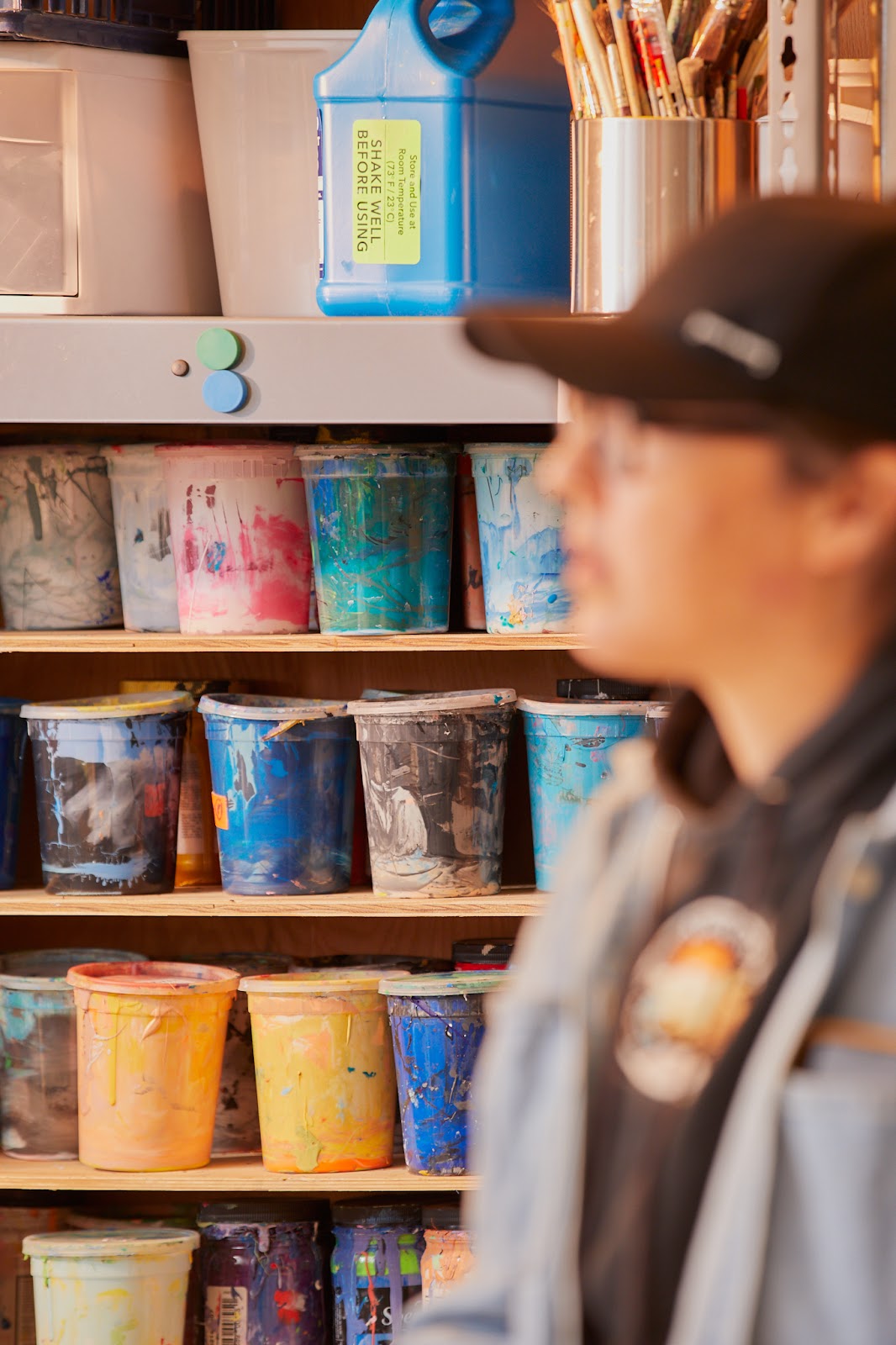 Image: Nadine Nakanishi, out-of-focus, facing the left. Behind her is a shelving unit with plastic tubs of colored liquids. On the top shelf, is a tin filled with paintbrushes and a large blue jug which says "SHAKE WELL BEFORE USING." Photo by Sarah Joyce. 