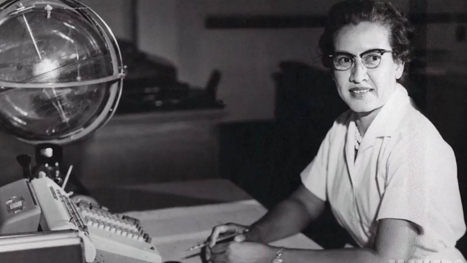 Black and white portrait of Katherine Johnson sitting at a desk with a white short sleeve polo and cat eyed glasses, smiling towards the camera. There is a computer and a glass globe-like instrument in front of her.