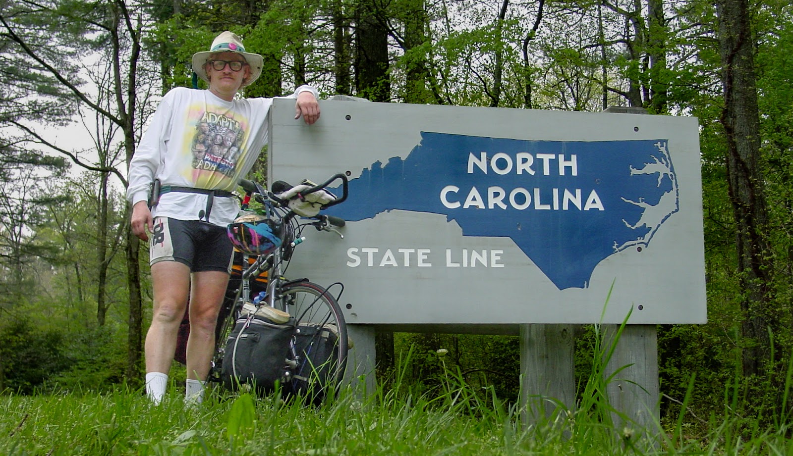 Cyclist in a wide-brimmed hat leans on the North Carolina state line marker. 