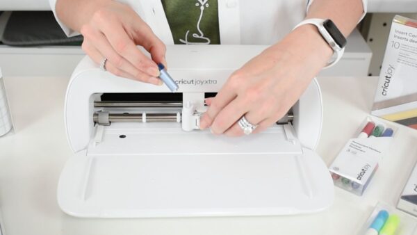 The image shows Abbi Kirsten's hands replacing the fine point blade with the foil tool in the Cricut Joy Xtra machine. 